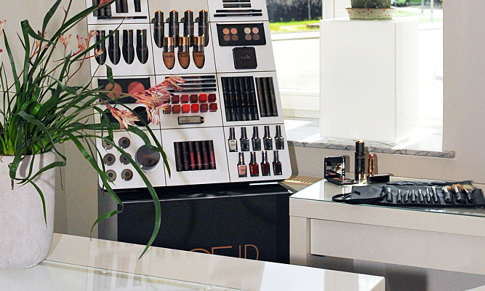 Make-up-Display mit Lippenstiften, Lidschatten und Nagellacken im Institut, daneben eine Pflanze
