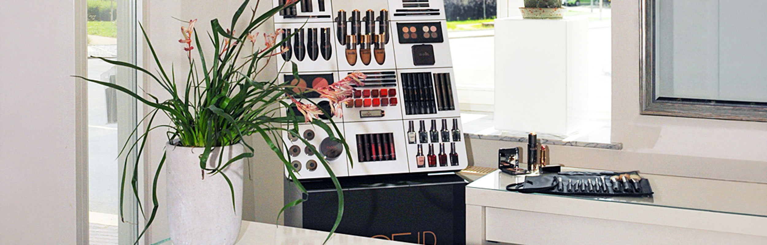 Make-up-Display mit Lippenstiften, Lidschatten und Nagellacken im Institut, daneben eine Pflanze